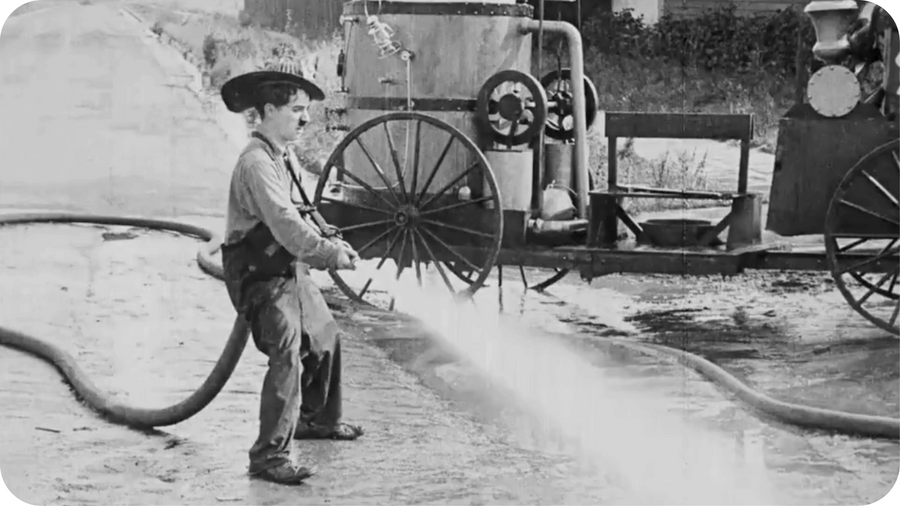 Photographie de Charlie Chaplin en train d'utiliser une lance de pompier. 